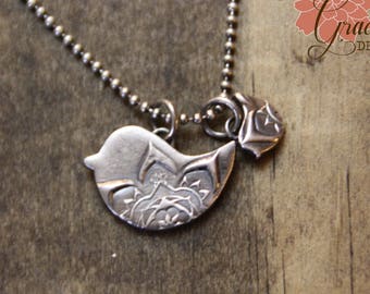 Puede incluir: Un collar de plata con un colgante de pájaro y un pequeño amuleto. El colgante de pájaro tiene un diseño detallado y el amuleto tiene forma de flor.