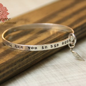 May include: Silver bangle bracelet with the inscription "God has you in his arms" and a small angel wing charm.