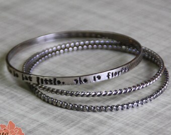 May include: Three silver bangles stacked on top of each other. The top bangle has the words "she is fierce but little" engraved on it.
