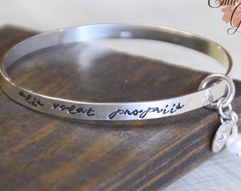 May include: Silver bangle bracelet with a charm and a pearl. The bracelet is engraved with the text "alit volat propriis".