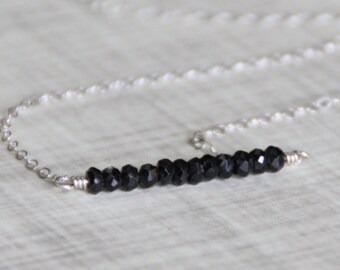 May include: A delicate silver necklace featuring a horizontal bar of small, faceted black beads. The beads are strung together and attached to a fine silver chain. The necklace is displayed on a light-colored surface.