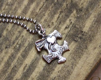 Puede incluir: Un collar de plata con un colgante de pieza de rompecabezas con un pequeño corazón en el centro. El collar está sobre una superficie de madera.