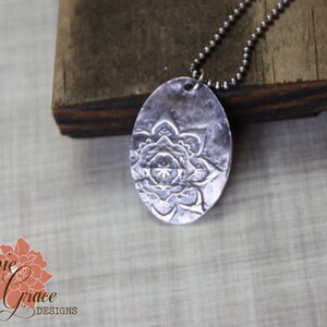 May include: A silver necklace with a large oval pendant featuring a detailed mandala design. The pendant is textured and has a hammered finish.