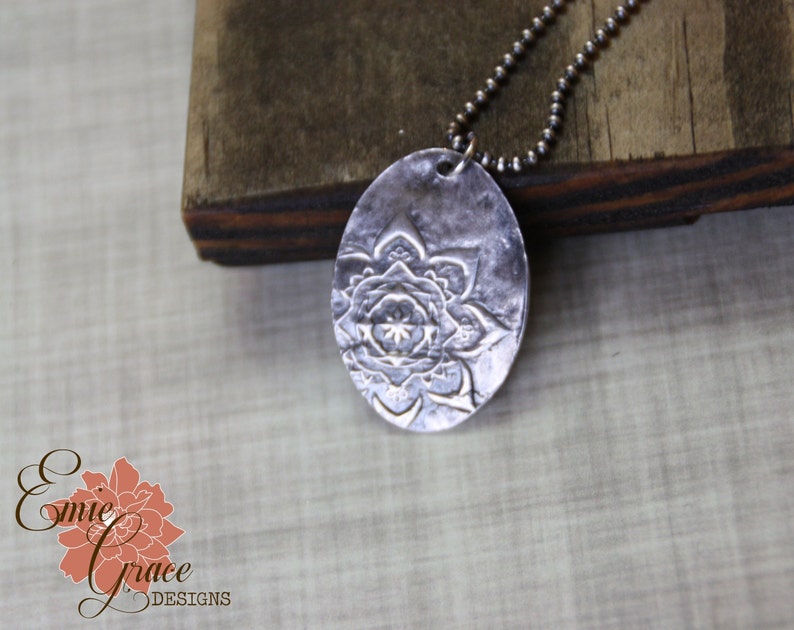 May include: A silver necklace with a large oval pendant featuring a detailed mandala design. The pendant is textured and has a hammered finish.