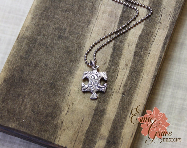 Puede incluir: Un collar de cadena de plata con un colgante de pieza de rompecabezas de plata. El colgante tiene un dise&ntilde;o detallado y texturizado.