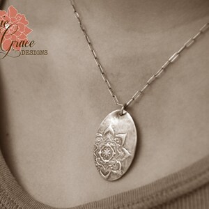 May include: A silver necklace with a pendant featuring a detailed mandala design. The pendant is oval shaped and hangs from a silver chain.