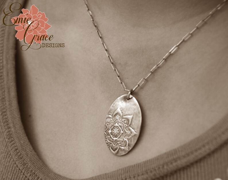 May include: A silver necklace with a pendant featuring a detailed mandala design. The pendant is oval shaped and hangs from a silver chain.