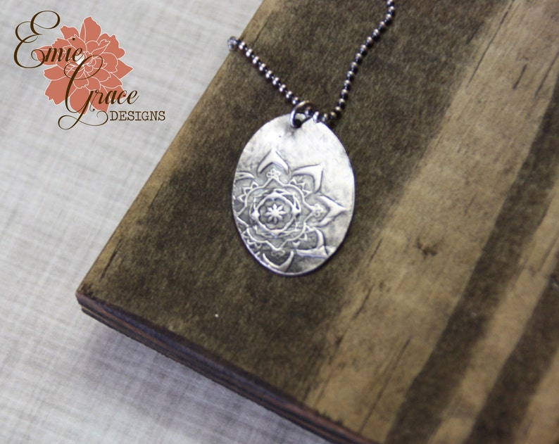 May include: A silver necklace with a round pendant featuring a detailed mandala design. The pendant is hanging on a silver chain.