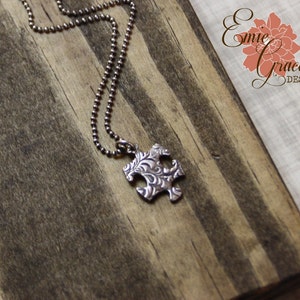 Puede incluir: Un collar de cadena de plata con un colgante de pieza de rompecabezas de plata. El colgante tiene un dise&ntilde;o floral detallado.