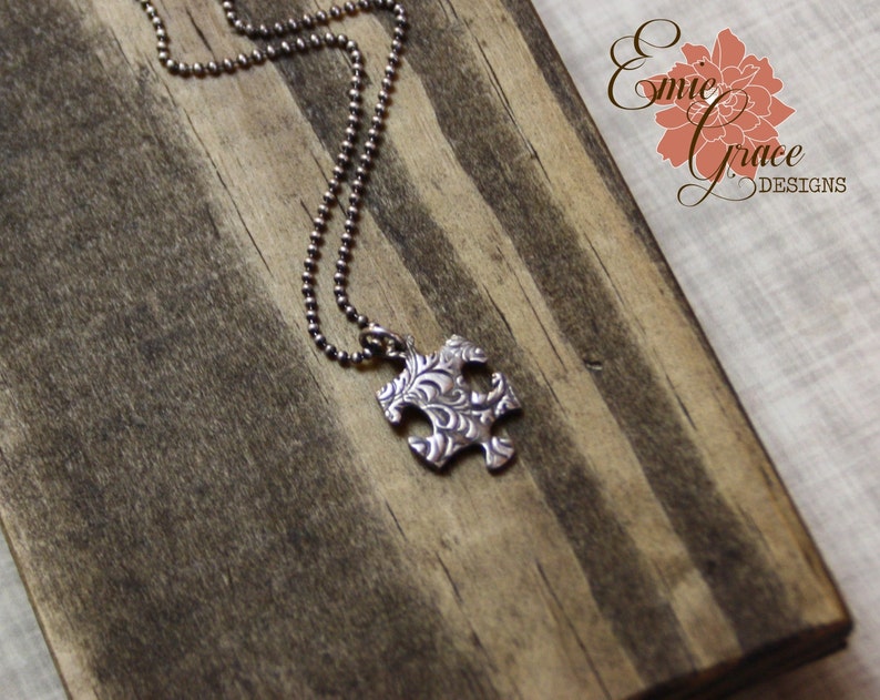 Puede incluir: Un collar de cadena de plata con un colgante de pieza de rompecabezas de plata. El colgante tiene un dise&ntilde;o floral detallado.