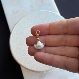 Könnte beinhalten: Ein kleiner, weißer Teekannen-Anhänger mit goldenen Akzenten und einem goldenen Verschluss. Der Anhänger wird in einer Hand vor einem weißen und schwarzen Hintergrund gehalten. Die Teekanne hat ein zartes Design, das auf ein Schmuck- oder Accessoire-Artikel hindeutet.