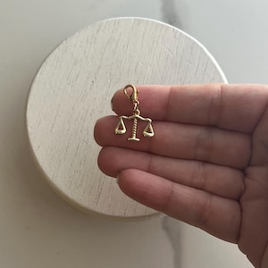 May include: A gold-coloured charm featuring the scales of justice. The charm is suspended from a small clasp, and the scales are balanced. The charm is held against a white background.