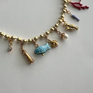 Op de afbeelding: Gouden ketting met maritieme bedels. De ketting heeft een vuurtoren, vissen, koraal en een kreeft. De ketting is van kralen en er is een parelbedel. De bedels zijn geschilderd in blauw, rood en goud. Een charmant accessoire.