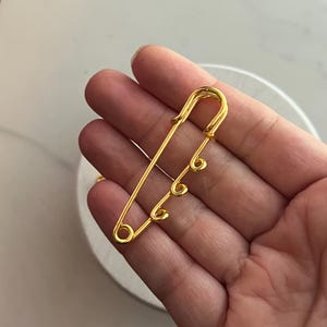 May include: A gold-colored safety pin with three decorative loops. The pin is a large size and has a classic design. The metal has a shiny finish. The pin is held in a hand.