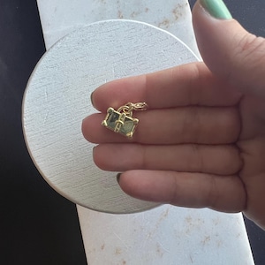 May include: A small, gold-colored charm with two square, green-tinted gemstones. The charm is held against a white and gray surface, with a black background. The charm is likely for a necklace or bracelet.