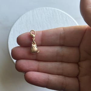 May include: A gold-colored cat charm with a lobster clasp. The charm features a small, rounded cat design with etched details. The charm is suspended from a gold-toned clasp, suitable for a bracelet or necklace. The charm is small and delicate.