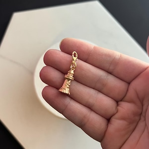 Può includere: Un ciondolo a forma di faro color oro con dettagli intricati, tra cui finestre e una lanterna. Il ciondolo è sospeso da una piccola chiusura, pronto per essere aggiunto a una collana o un braccialetto. Il ciondolo è tenuto in mano.