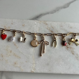 Op de afbeelding: Gouden bedelarmband met diverse bedels, waaronder een rood hart, parel, vlinder, strik, wensbeen, lieveheersbeestje en klavertjevier. De armband wordt getoond op een wit marmeren oppervlak.