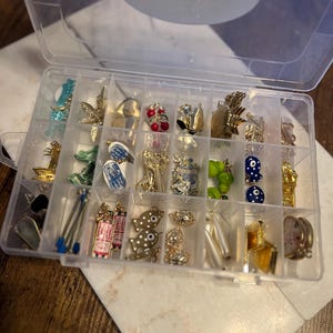 May include: A clear plastic organizer filled with various small charms. The charms include gold-colored figurines, cherries, and blue and white designs. The organizer sits on a white and gray marble surface.