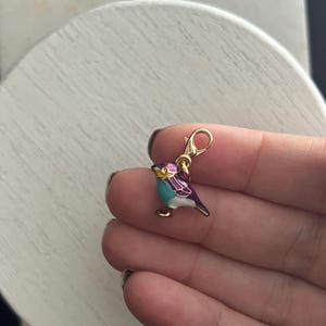 Puede incluir: Un charm de oro con un diseño de pájaro azul y morado. El pájaro tiene un pico y patas doradas.
