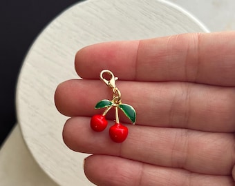 May include: Gold charm featuring two red cherries with green leaves. The charm has a lobster clasp.