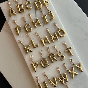 May include: A collection of gold-colored alphabet charms, each with a lobster clasp for easy attachment. The letters are in a bold, rounded font, and are displayed on a white and gray marble surface. The charms are arranged in alphabetical order.