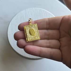 Puede incluir: Un medallón dorado en forma de libro con una abertura ovalada en la parte delantera. El medallón tiene detalles intrincados y un cierre en el lateral.