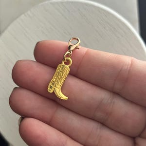May include: Gold charm in the shape of a cowboy boot. The charm has a detailed design and a lobster clasp.