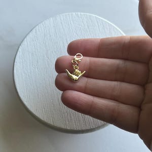 May include: Gold charm in the shape of a hand making the shaka sign, a popular Hawaiian gesture. The charm has a lobster clasp for attaching to a bracelet or necklace.
