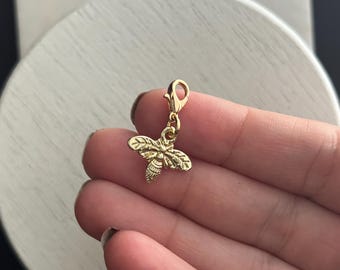 May include: A gold-coloured bee charm with detailed wings and body segments, attached to a clasp. This charm is designed to be added to a bracelet or necklace. The bee is a symbol of nature and industry.