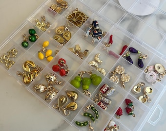 May include: A clear plastic organizer filled with various gold-toned and colorful charms. The charms include food items like avocados, lemons, and strawberries, as well as other designs. The organizer is on a white marble surface.