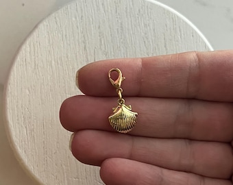 Op de afbeelding: Een goudkleurige schelpenbedel met een karabijnsluiting. De bedel is gedetailleerd met geribbelde lijnen en een kleine lus voor bevestiging. De bedel is ontworpen om aan een armband of ketting te worden bevestigd.