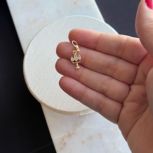 Könnte beinhalten: Ein kleiner, goldfarbener Anhänger mit einem Kronleuchter-Design. Der Anhänger hat klare, rechteckige und quadratische Kristalle. Der Anhänger ist an einer kleinen Schlaufe aufgehängt. Der Anhänger wird in der Hand einer Person gehalten.