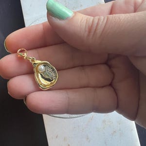 May include: A gold-colored charm with a pearl and small crystals. The charm is in the shape of an oyster shell. The charm is held by a person with light green nail polish.
