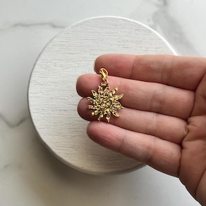 May include: A gold-toned sun charm featuring a textured, uneven surface and multiple radiating points. The charm includes a small lobster claw clasp at the top, allowing it to be easily attached to various items like bracelets or necklaces.