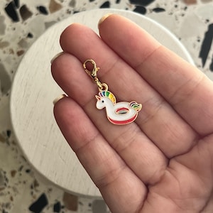 Könnte beinhalten: Ein goldfarbener Anhänger mit einem weißen Einhorn mit Regenbogenmähne und -schwanz. Der Anhänger hat rote und grüne Akzente und ist an einem goldfarbenen Verschluss befestigt. Der Anhänger wird in einer Hand gehalten.