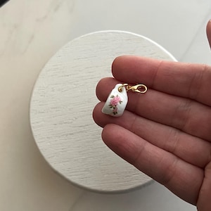 May include: A small, white porcelain charm with a gold-colored clasp. The charm features a delicate floral design in pink and green. The charm is held in a hand against a white background.