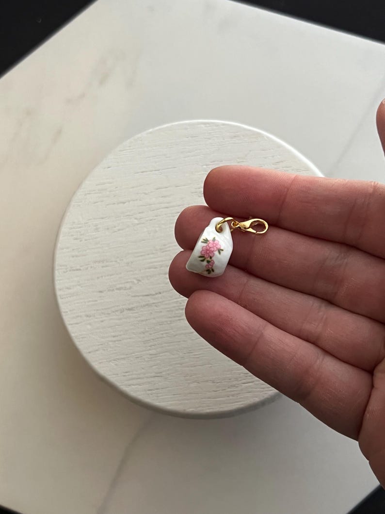 May include: A small, white porcelain charm with a gold-colored clasp. The charm features a delicate floral design in pink and green. The charm is held in a hand against a white background.