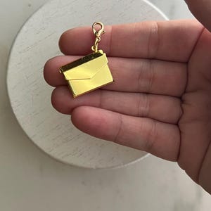 May include: A gold-colored charm in the shape of an envelope, with a clasp at the top. The charm is shiny and reflective, with a simple, elegant design. The charm is likely for a bracelet or necklace.