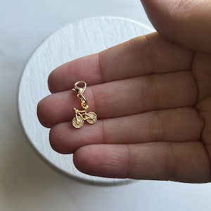 Puede incluir: Un charm de bicicleta de color dorado con detalles claros y brillantes en las ruedas. El charm está unido a un cierre, listo para ser añadido a una pulsera o collar. El charm es pequeño y delicado, ideal para joyería.