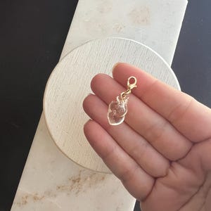 Puede incluir: Un dije transparente en forma de corazón con un cierre dorado. El dije se sostiene en una mano sobre un fondo de superficie de mármol blanco y fondo negro. El dije parece estar hecho de vidrio o un material transparente similar.