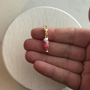 May include: A gold-coloured charm in the shape of a pink milkshake with whipped cream and a cherry on top. The charm is attached to a gold clasp. The charm is held in a hand.