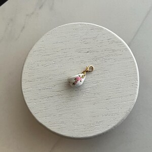 May include: A miniature white teacup charm with a gold clasp. The teacup features a delicate pink floral pattern. The charm is small and detailed, perfect for jewelry making or as a decorative accent.