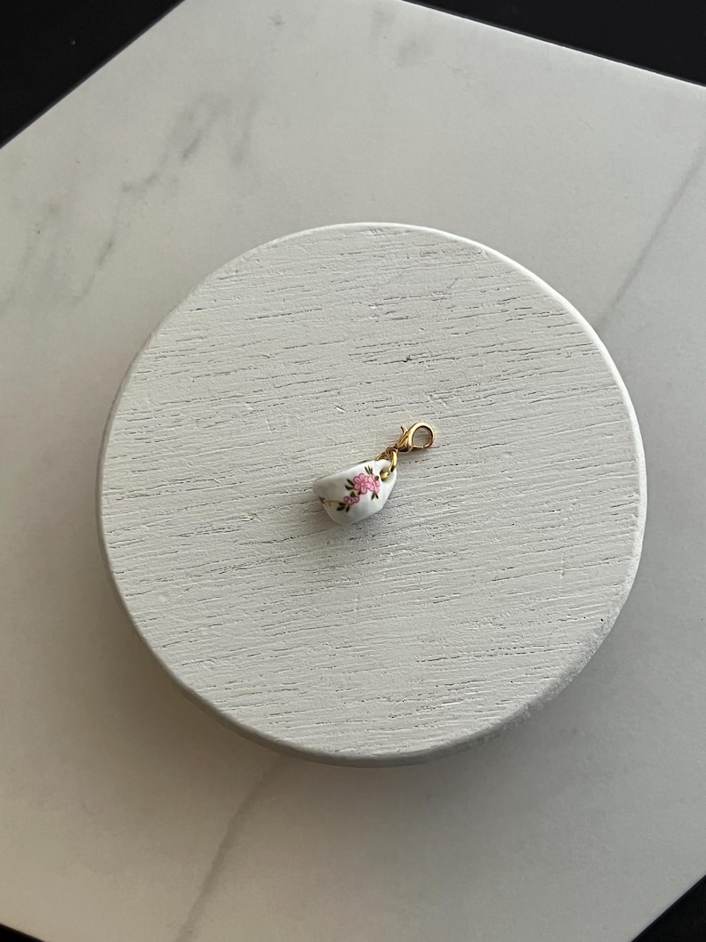 May include: A miniature white teacup charm with a gold clasp. The teacup features a delicate pink floral pattern. The charm is small and detailed, perfect for jewelry making or as a decorative accent.