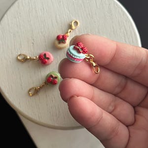 Puede incluir: Una colección de dijes de macaron en miniatura en colores pastel, incluyendo rosa, azul y verde, cada uno adornado con una cobertura de bayas rojas. Los dijes tienen cierres dorados, perfectos para la creación de joyas o proyectos de artesanía.