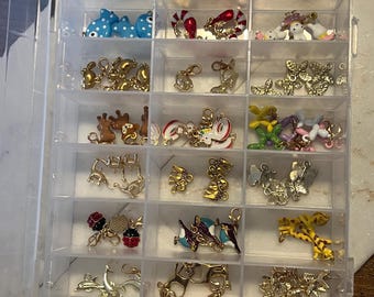 May include: A clear plastic organizer filled with various small charms. The charms include animals like giraffes, fish, and unicorns, along with other decorative elements. The charms are in gold, blue, red, and other colors.