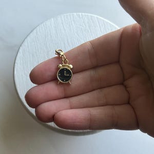 Puede incluir: Encanto de reloj despertador dorado con una esfera negra. El reloj tiene una pequeña aguja negra que apunta a las 12 y una aguja negra más grande que apunta a las 3.