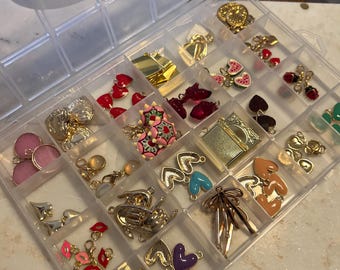May include: A clear plastic organizer filled with various jewelry charms. The charms include gold-toned bows, hearts, and other shapes, along with red, pink, and blue accents. The organizer is on a white marble surface.