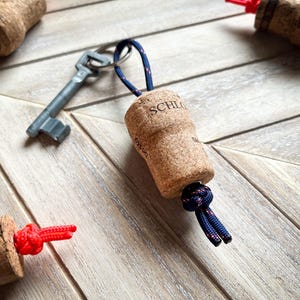 May include: A cork keychain with a blue and red paracord loop. The cork is engraved with the word "SCHL" and is attached to a silver key.