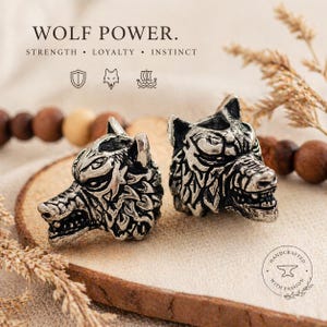 Könnte beinhalten: Zwei silberfarbene Wolfskopfperlen mit aufwendigen Details, präsentiert auf einer Holzoberfläche. Das Bild enthält den Text "WOLF POWER" und die Worte "STRENGTH, LOYALTY, INSTINCT". Die Perlen sind handgefertigt mit Leidenschaft.
